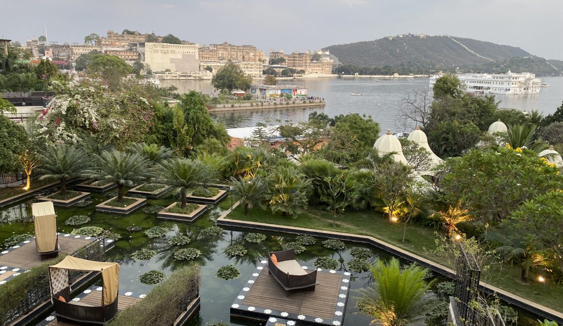 This image shows the gardens of the Leela Palace Udaipur with the City Palace and Lake PIchola in the background